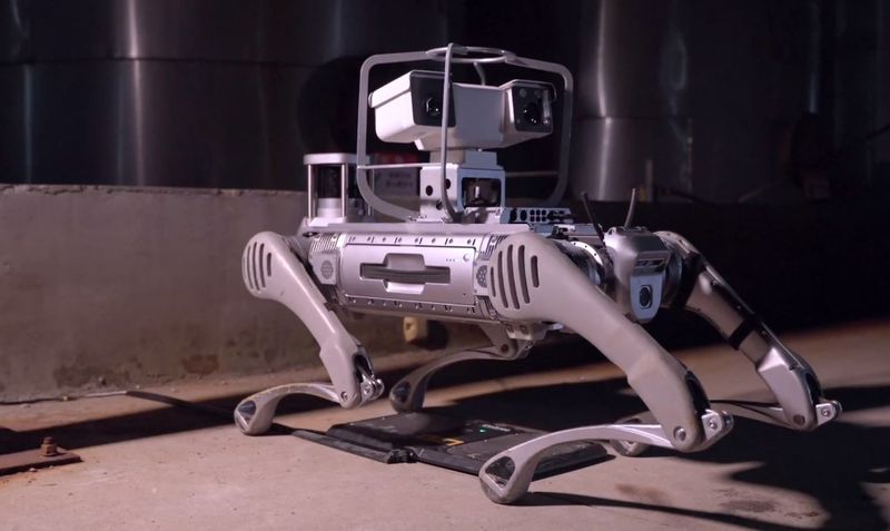 Unitree B2 with a pan-tilt inspection camera payload standing on its charging station in an industrial plant